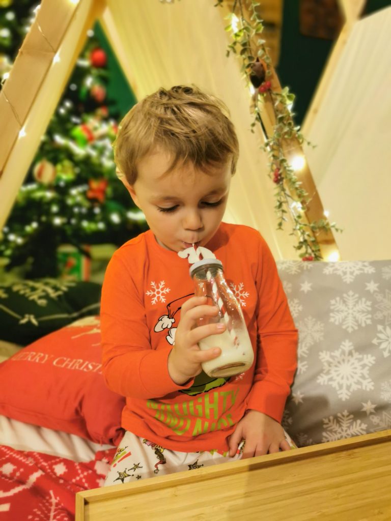 Small child in red pyjamas drinking milk from glass bottle with a straw. Fairy lights glow behind him.