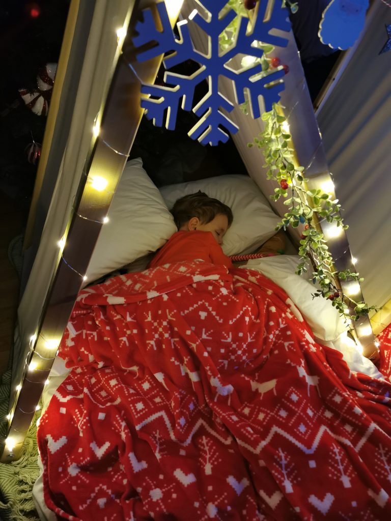 Small child with red blanket over them asleep in little teepee. Fairy lights at the entrance.
