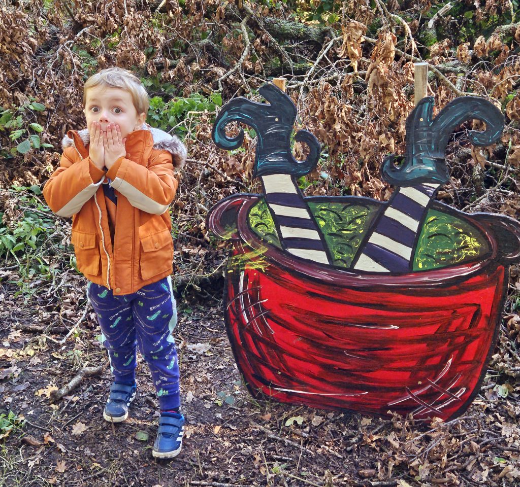 Child with hands over mouth stood next to cauldron with witches legs poking out  