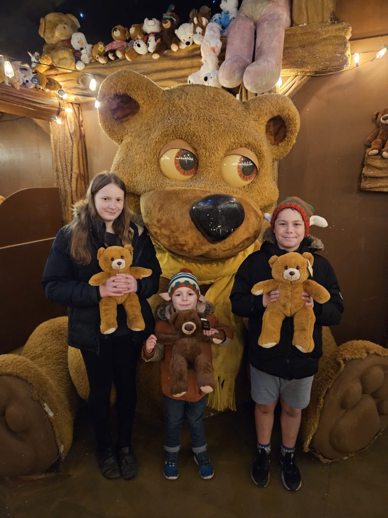 3 children holding teddy bears stood in front of giant teddy bear