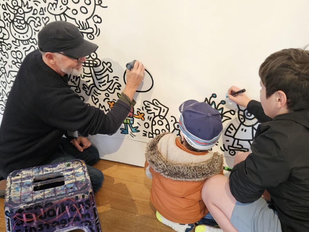 Artist Scott Walker drawing doodles on the gallery wall with 2 children next to him